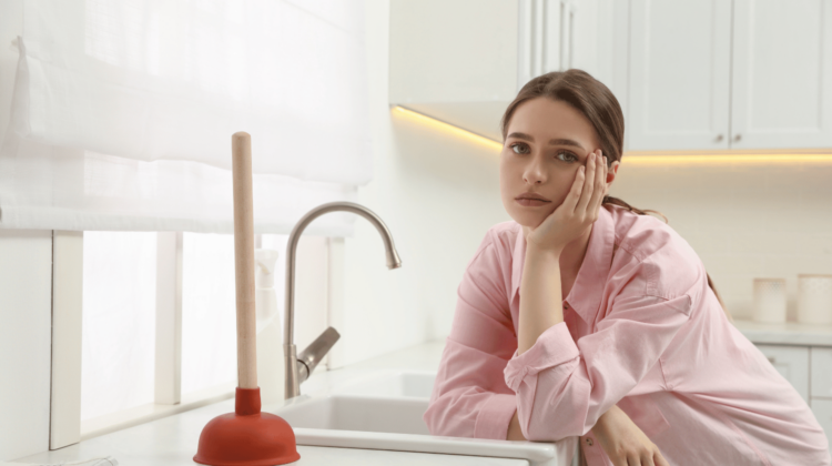 Frustrated woman with clogged kitchen sink and plunger on countertop