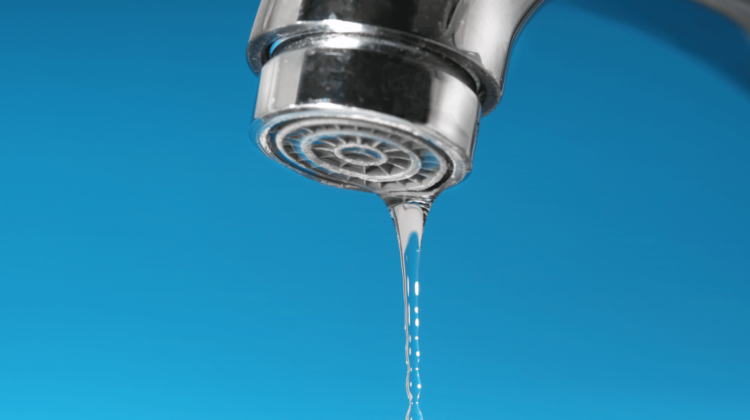 Extreme close-up of a modern chrome faucet dripping water against a bright blue background, symbolizing a plumbing leak and water waste.